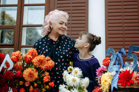 A beautiful elderly grandmother smiles at her cute granddaughter with a cool hairstyle. They hug. In front of them are baskets with large bright flowers. Take a walk in the fresh air. Tenderness.の写真素材