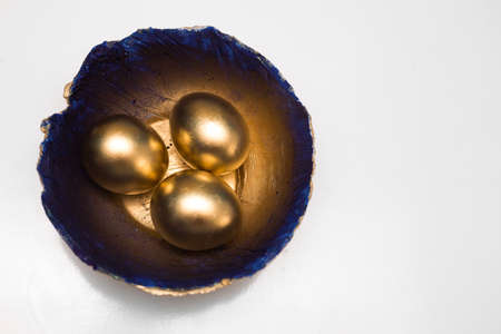Photo of 3 golden Easter eggs lying on a decorative handmade plate painted in blue and gold. Top view, white background. Stylish and unusual DIY Easter home decor.の写真素材