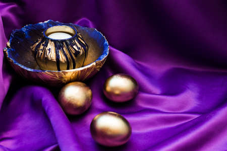 Decorative Easter set of a candlestick painted in gold and blue and plates under it. Nearby lie 3 golden eggs, the composition stands on a purple satin flowing background. Festive decorationの写真素材