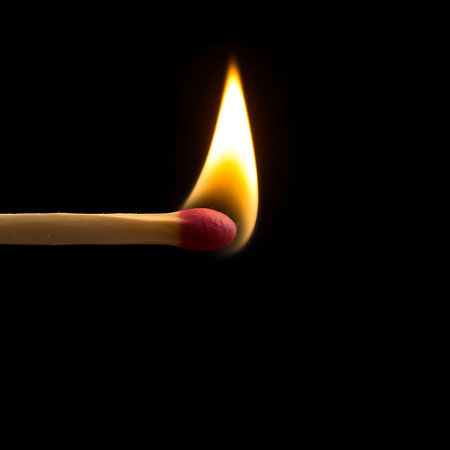 A single matchstick with a red tip ignites with a bright orange flame against a stark black background, symbolizing fire and lightの素材