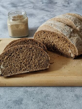 cut rye bread on a wooden plank and a jar of sourdough against the background on gray table. High...