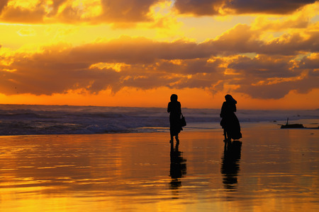 Two women walking along the ocean shore during sunsetの写真素材