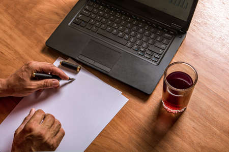 Hands of a man writing with ink pen on white paper. Laptop keyboard, glass of juice.