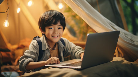Happy asian boy using laptop computer while sitting in tent at homeの素材