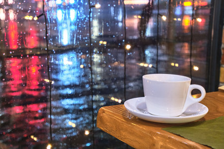 Coffee cup on the edge of a table near a window with rain outside.の写真素材