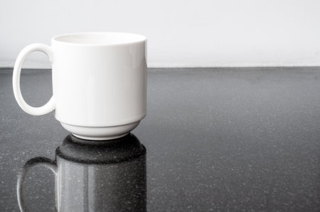 White coffee cup on black granite table with white wall and reflection.