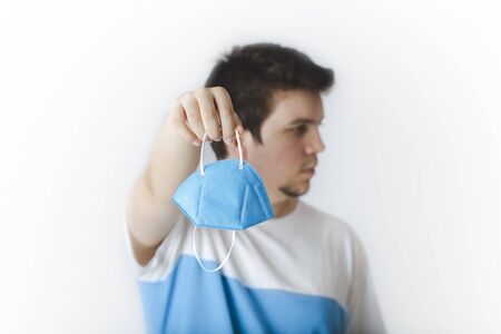 EUROPEAN CAUCASIAN YOUNG MAN IN WHITE BACKGROUND HOLDING A FFP2 BLUE FACE MASK PREVENTION DURING QUARANTINE TO AVOID THE SPREADING OF COVID-19 CORONAVIRUSの写真素材