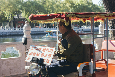 25,Oct,2014 -Beijing,China. side profile of Man on rickshaws at Houhai Lake. Tourists can have  a rickshaw tour around Houhai Lake to explore the ancient hutongs and traditional building. the board show  the name of places he can goのeditorial素材