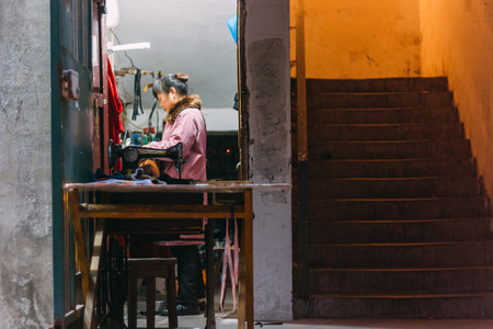 Chongqing, CHINA - 22, Dec, 2015: A chinese women  tailor entrepreneur sewing behind an old machine in a street beside stairsのeditorial素材