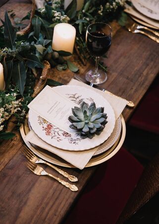 Rustic wedding table seting with vintage plates, green and white eucalyptus garland, deer antlers, gold utensils. Boho style. Table set for an event, party, date or wedding. Red wine servedの写真素材