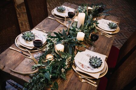 Rustic wedding table seting with vintage plates, green and white eucalyptus garland, deer antlers, gold utensils. Boho style. Table set for an event, party, date or wedding. Red wine servedの写真素材