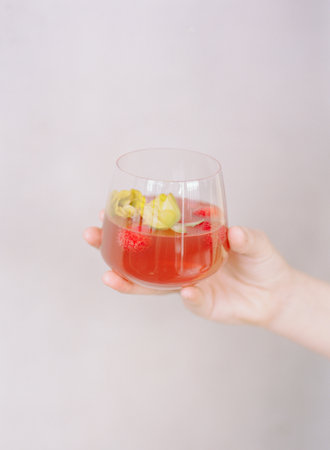 A hand holds a clear glass filled with a vibrant red cocktail, garnished with edible flowers and...