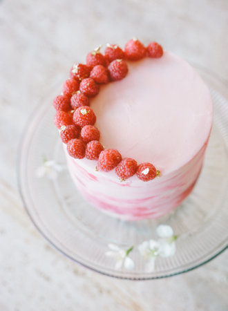 A round cake adorned with fresh raspberries and delicate flowers is displayed on a glass stand, set...
