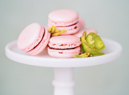 Delicate pink macarons displayed on a white cake stand with green flowers in a softly lit settingの写真素材