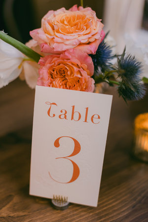 At a wedding reception, a table number display features a floral arrangement of pink and orange roses, elegantly marking Table 3 among warm lighting.の写真素材