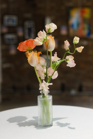 A delicate arrangement of orange and pink flowers stands elegantly in a clear glass vase on a round white table. The background features colorful artwork and a warm ambiance.の写真素材