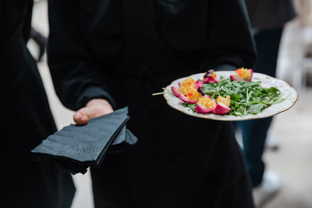 A chef in a dark uniform offers a plate featuring a vibrant salad adorned with colorful edible flowers, accompanied by neatly stacked black napkins. The setting suggests an elegant gathering.の写真素材