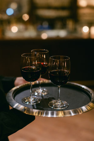 Waitstaff carries a silver tray with three glasses of red wine in a dimly lit bar filled with warm lights.の写真素材