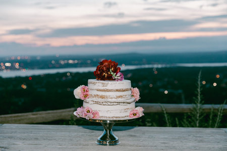 A beautifully decorated wedding cake with floral embellishments sits on a table, showcasing a stunning sunset view.の写真素材