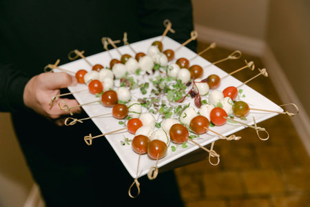 A platter filled with skewers of mozzarella balls and cherry tomatoes garnished with herbs is served at a gathering.の写真素材