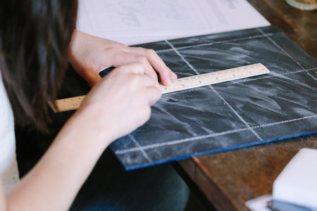 A person carefully uses a ruler to draw precise lines on a textured surface, sitting at a rustic...