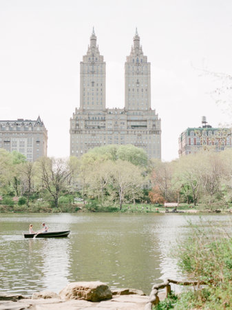 Two people paddle leisurely in a small boat on a tranquil lake surrounded by spring foliage and...