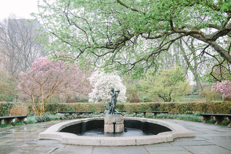 Spring colors burst in a serene garden featuring a stone fountain surrounded by vibrant flowering trees.の写真素材