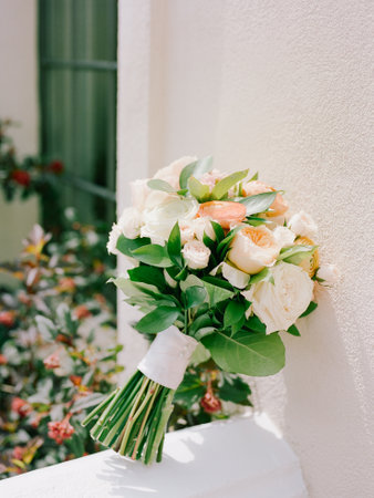 A stunning bouquet of fresh flowers leans against a white wall, surrounded by blooming greenery on a sunny day.の写真素材