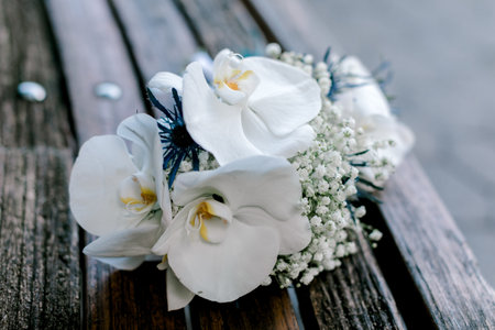 A delicate bouquet of white orchids and babyâ€™s breath rests on a weathered wooden surface,...