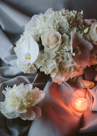 A beautiful bouquet of white flowers arranged on soft fabric, accompanied by a lit candle creating a serene atmosphere.の写真素材