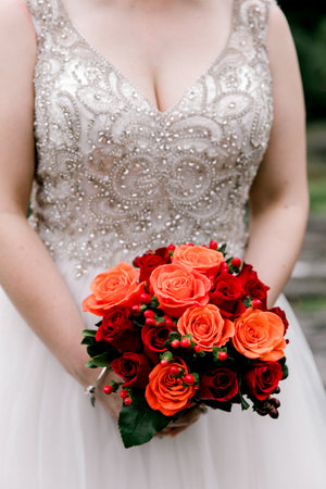 A happy bride shows off a stunning red and orange rose bouquet in a beaded gown at a serene outdoor venue.の写真素材