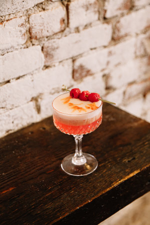 A vibrant raspberry cocktail garnished with fresh raspberries sits on a wooden table against a rustic brick wall.の写真素材