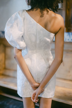 Model stands confidently, showcasing a stylish one-shoulder dress with a dramatic sleeve in a warm, inviting room.の写真素材