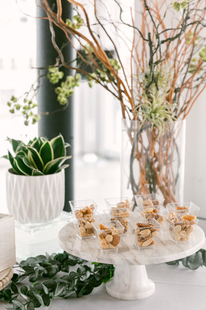 Small, clear cups filled with assorted snacks sit on a marble stand surrounded by greenery and decorative branches.の写真素材