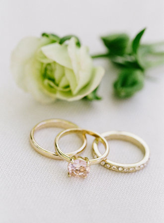 Delicate gold rings, one with a pink gem, arranged beside soft flowers on a white surface at a...