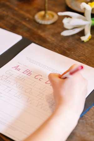 A child is focused on writing letters in a workbook using a red marker while surrounded by flowers and natural light.の写真素材