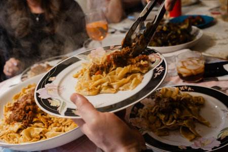 Steam rises from a plate of hearty pasta as a person serves a generous portion at a lively family dinner.の写真素材