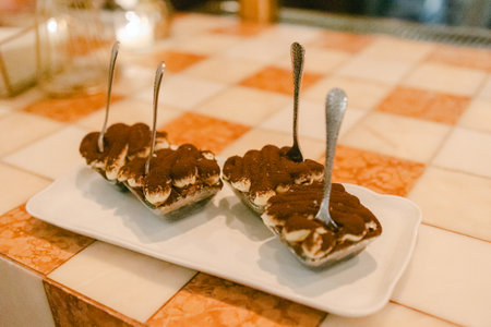Three servings of tiramisu with spoons displayed on a patterned table in a warm-lit restaurant setting.の写真素材