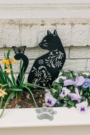 A black metal cat silhouette stands beside colorful flowers and a butterfly in a spring garden.の写真素材
