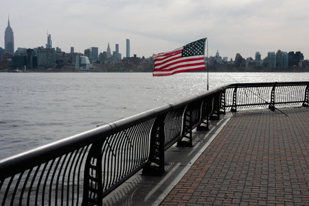 An American flag waves gently by the waterfront as the New York City skyline looms in the background under cloudy skies.の写真素材