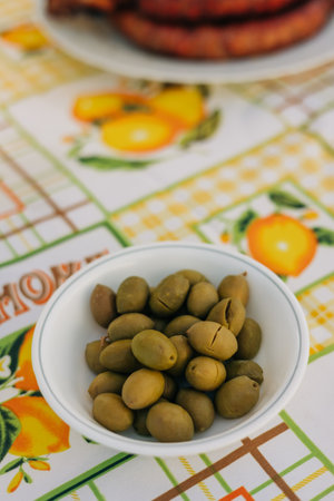 A small bowl filled with green olives sits on a colorful tablecloth decorated with fruit patterns during a relaxed meal.の写真素材