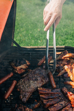 A person uses tongs to flip assorted grilled meats and vegetables at a lively backyard barbecue on a sunny day.の写真素材