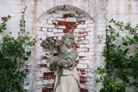 A womanâs statue carries a basket of flowers amidst greenery and rustic brick, creating a serene garden atmosphere.の写真素材