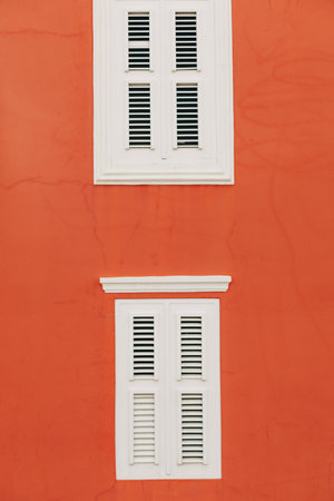 Two sets of white louvered windows stand out against a vibrant orange wall during a sunny day.