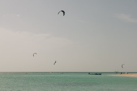 Enthusiastic kite surfers glide over clear waters while boats drift nearby on a calm and sunny tropical beach.の写真素材