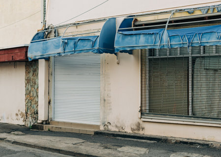 A shopfront featuring a blue awning and closed shutter on a calm street at midday, with a weathered appearance.の写真素材