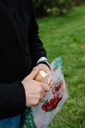 Individual uses a knife to peel an apple while standing on fresh grass surrounded by trees in...