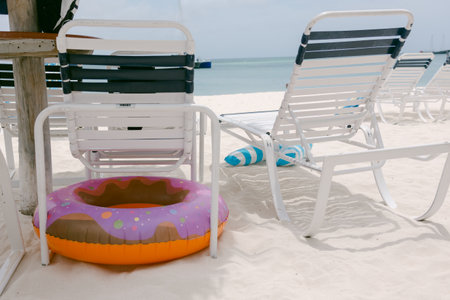 Bright pool float sits next to white lounge chairs on sandy beach, surrounded by calm turquoise...