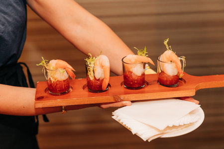 A person presents a wooden platter with shrimp cocktails in glasses during a lively gathering.