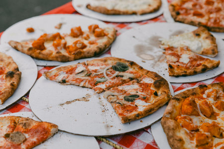 Various pizza slices rest on paper plates at a bustling outdoor event during a warm afternoon.の写真素材